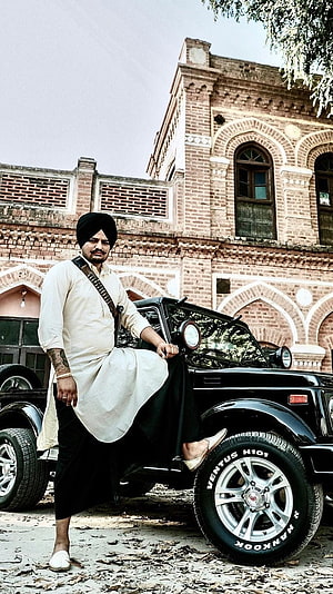 Sikh man in traditional attire posing with a black SUV in front of a historic brick building, Sidhu Moose Wala, Black Jeep, Background HD phone wallpaper