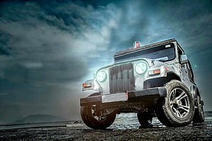 Silver Jeep Wrangler with glowing headlights parked on a wet beach under a dramatic cloudy sky HD wallpaper