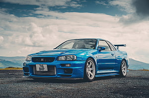 Blue Nissan Skyline GT-R R34 Coupe with Rear Spoiler on Gravel Road Under Cloudy Sky HD wallpaper