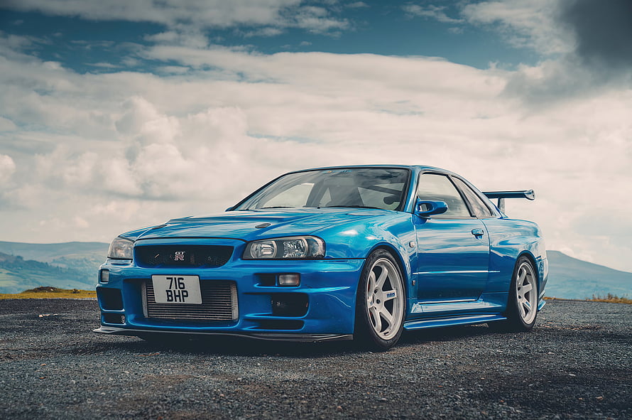 Blue Nissan Skyline GT-R R34 Coupe with Rear Spoiler on Gravel Road Under Cloudy Sky HD wallpaper