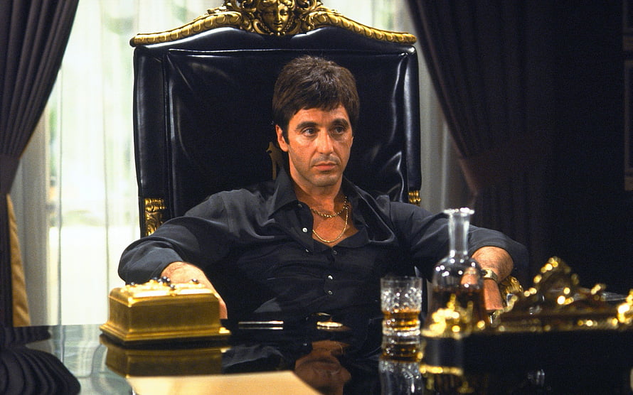 Tony Montana sitting in a black leather chair with gold accents in Scarface movie scene HD wallpaper