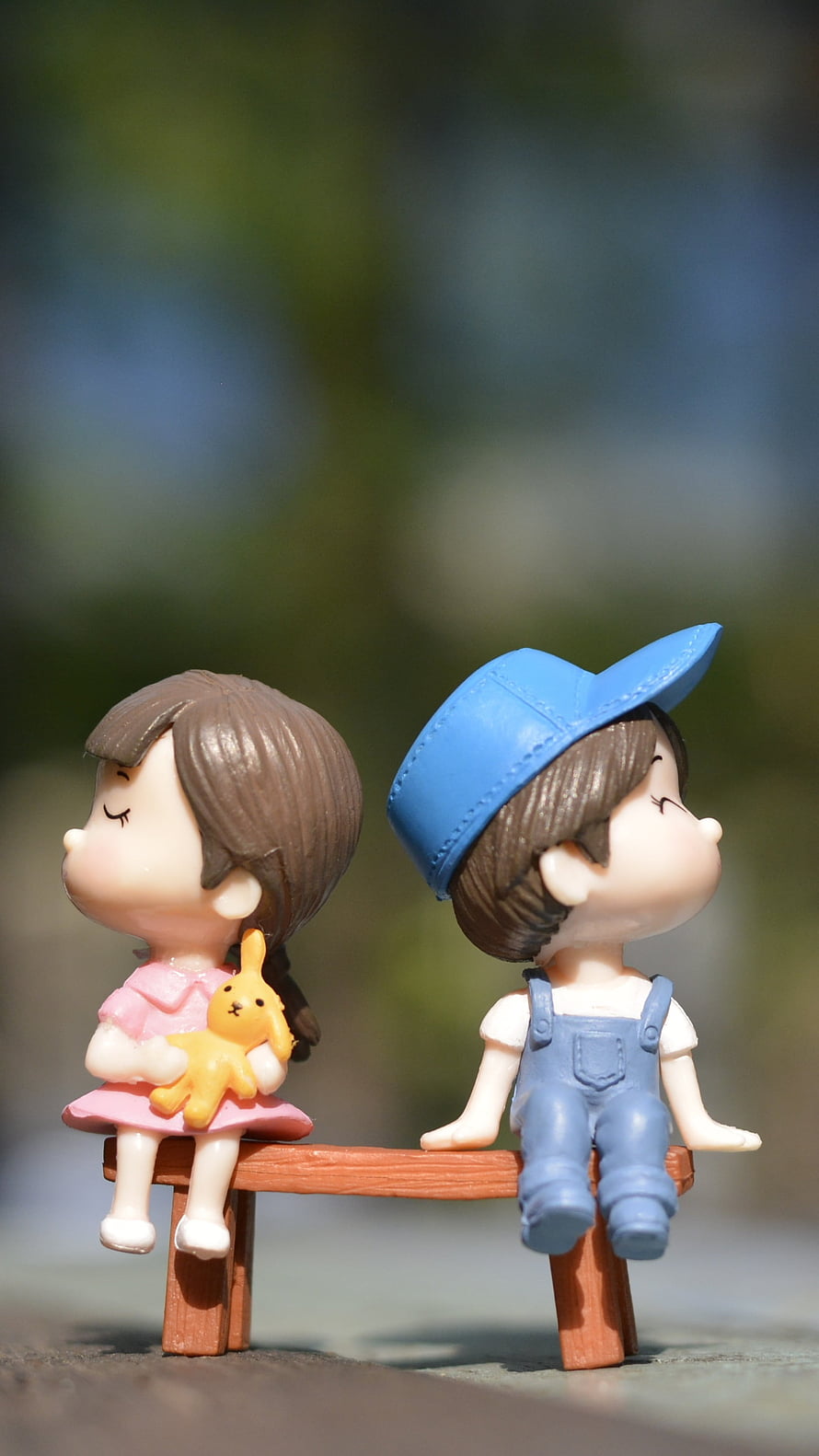 Cute Chibi Couple Figurines Sitting on a Miniature Bench with Soft Outdoor Background HD phone wallpaper