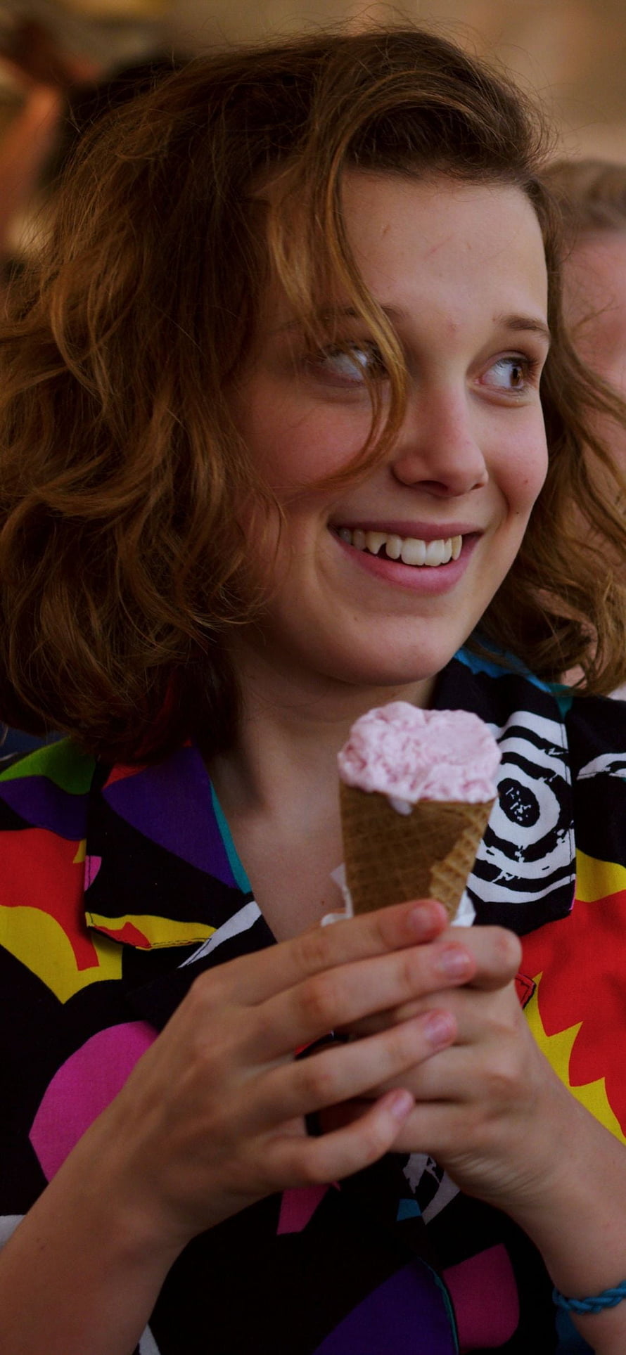 Millie Bobby Brown Smiling While Enjoying a Pink Ice Cream Cone in a Colorful Patterned Top HD phone wallpaper