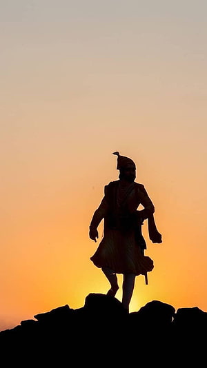 Silhouette of a historical warrior statue standing on a rocky peak against a vibrant orange sunset sky HD phone wallpaper