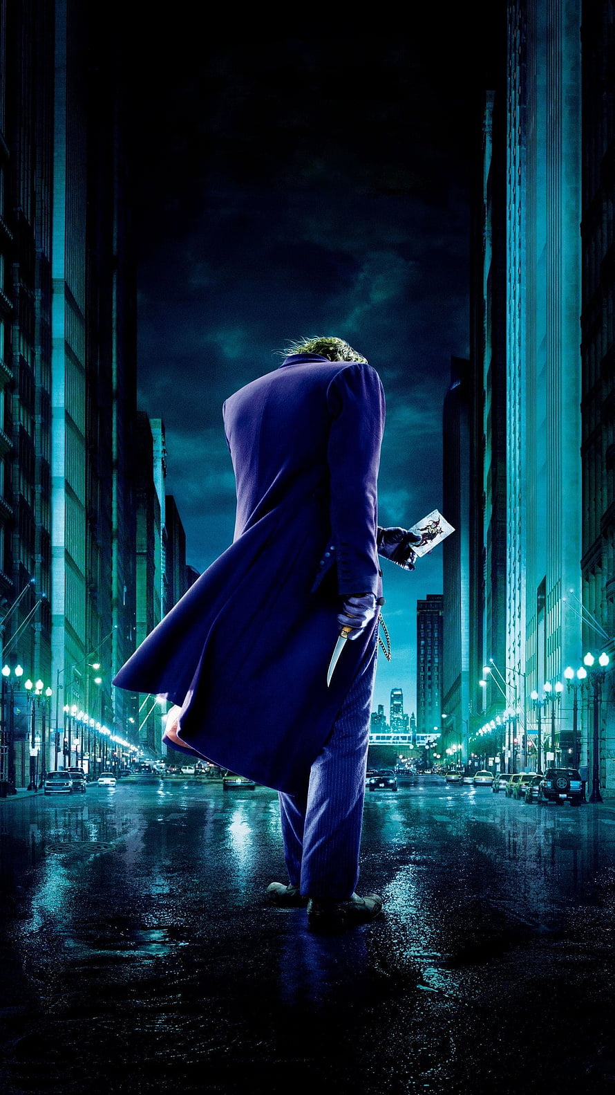 The Joker in a purple suit standing on a rainy city street at night holding a knife and a playing card HD phone wallpaper