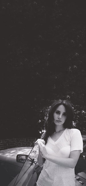 Lana Del Rey in a moody black and white portrait leaning against a car under dark trees HD phone wallpaper