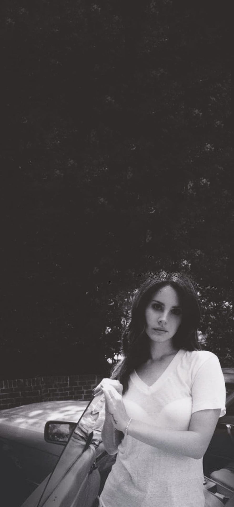 Lana Del Rey in a moody black and white portrait leaning against a car under dark trees HD phone wallpaper