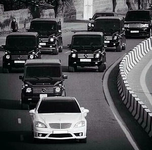 Convoy of Black Mercedes G-Class SUVs Following a White Sedan on a Curved Road HD wallpaper