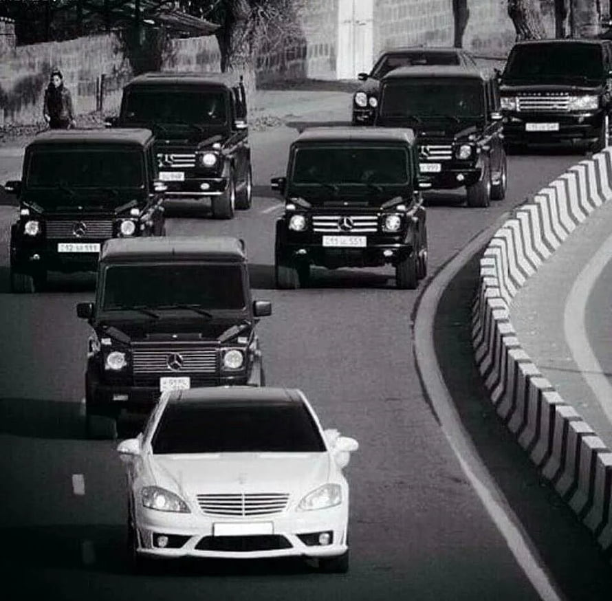 Convoy of Black Mercedes G-Class SUVs Following a White Sedan on a Curved Road HD wallpaper