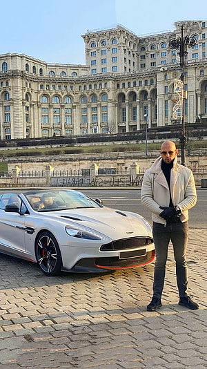 Andrew Tate standing beside a white Aston Martin DBS Superleggera in front of the Palace of the Parliament in Bucharest HD phone wallpaper