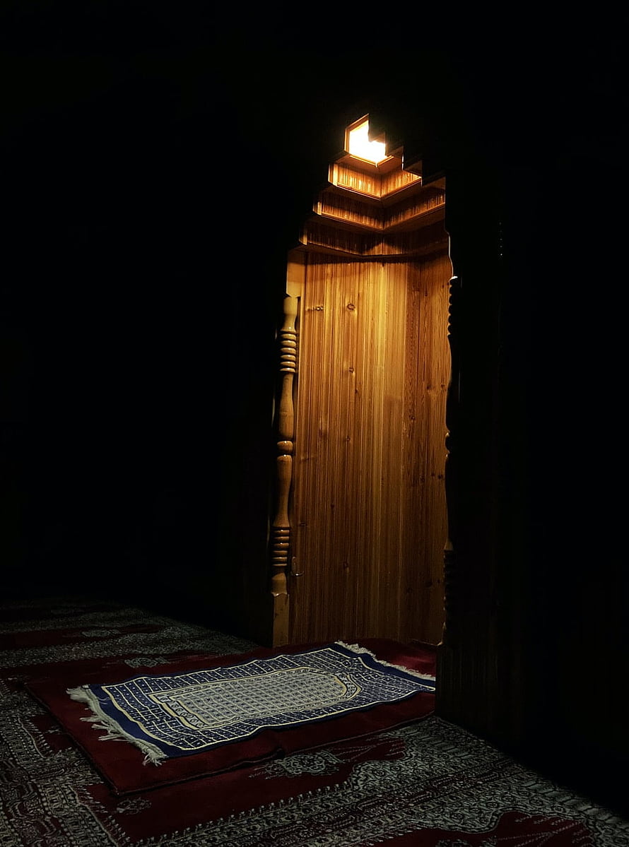 Illuminated wooden mihrab niche with prayer rug in dark mosque interior HD phone wallpaper