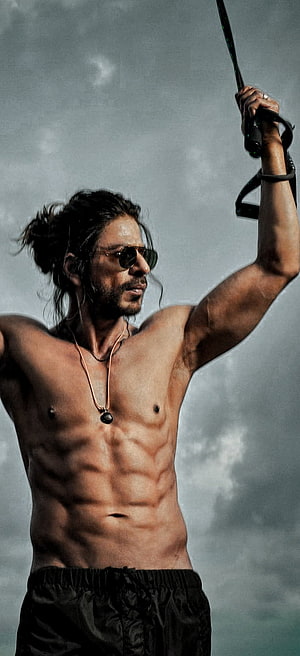 Shah Rukh Khan shirtless with sunglasses holding a sword against a cloudy sky HD phone wallpaper