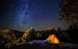 Illuminated orange tent glowing under a dense starry night sky in a mountainous landscape HD wallpaper