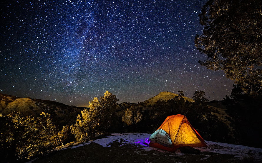 Illuminated orange tent glowing under a dense starry night sky in a mountainous landscape HD wallpaper