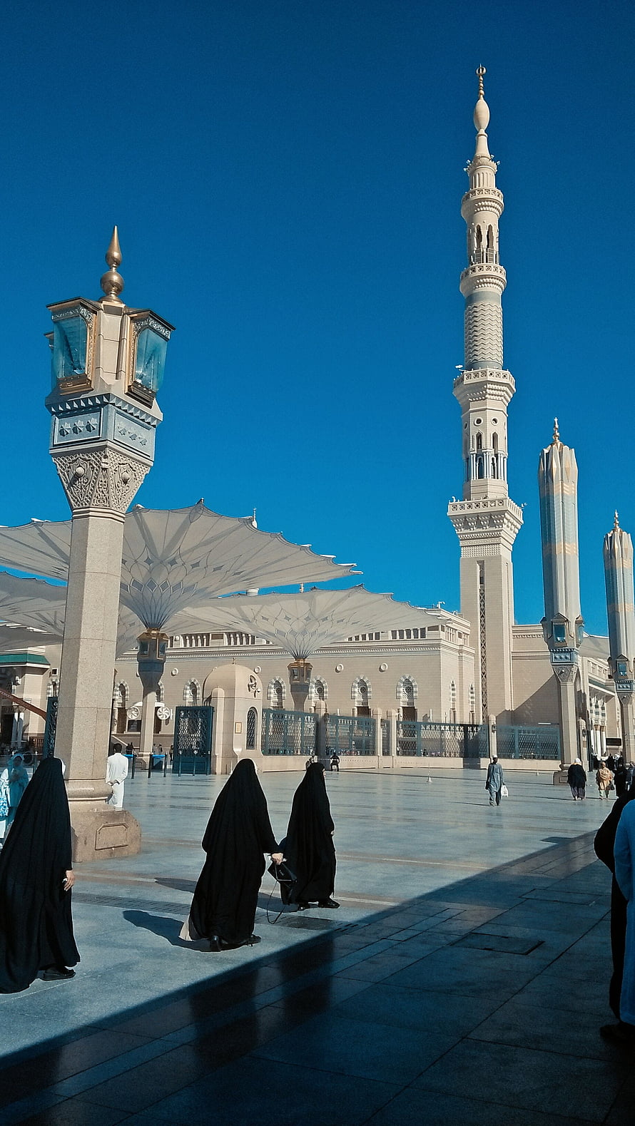 Nabawi Mosque courtyard with towering minarets and women in black abayas under a clear blue sky HD phone wallpaper