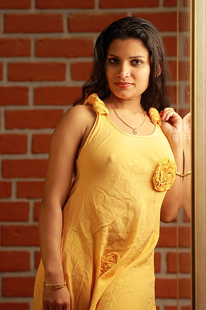 Reshmi Nair in a yellow sleeveless dress with floral details against a red brick wall HD phone wallpaper