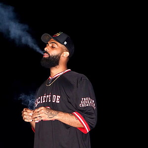 Drake exhaling smoke while wearing a black baseball cap and jersey against a dark background HD phone wallpaper