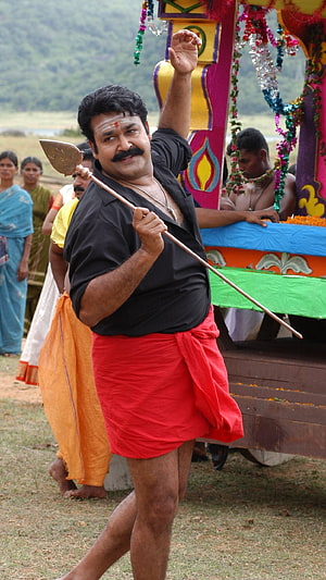 Mohanlal in traditional Kerala attire holding a ceremonial spear at a cultural festival HD phone wallpaper