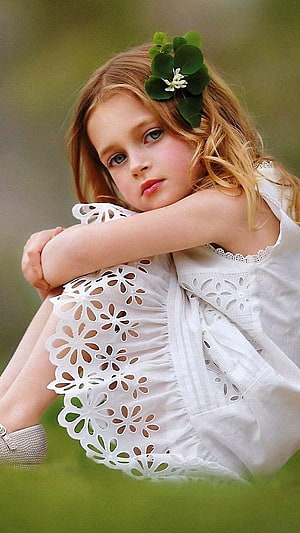 Portrait of a young girl with blue eyes wearing a white eyelet dress and a clover hair accessory sitting in a grassy field HD phone wallpaper