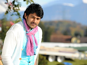 Prabhas in a white jacket and blue shirt with a pink scarf, smiling outdoors with a scenic background HD wallpaper