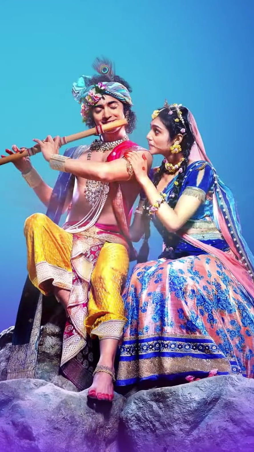 Radha Krishna playing flute on rock with peacock feather crown and traditional attire HD phone wallpaper