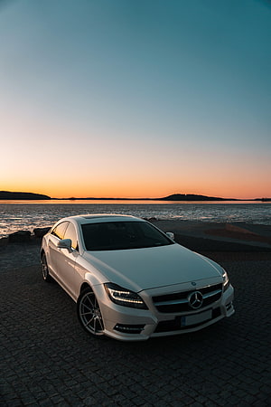 White Mercedes-Benz CLS parked on a cobblestone shore at sunset with a calm lake and distant hills in the background HD phone wallpaper