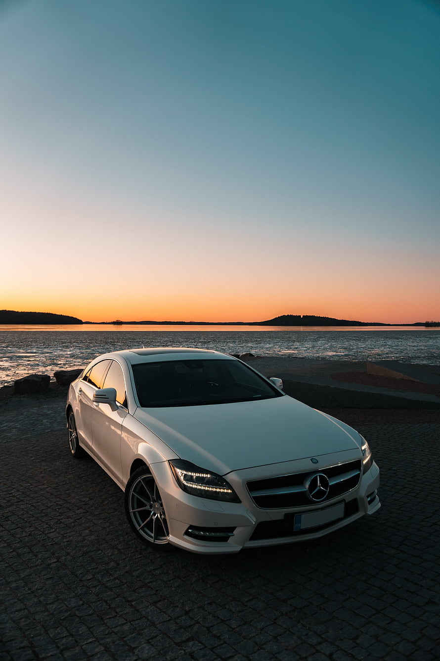 White Mercedes-Benz CLS parked on a cobblestone shore at sunset with a calm lake and distant hills in the background HD phone wallpaper