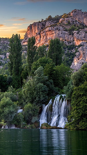 Serene waterfall cascading into a calm lake surrounded by lush green trees and rocky cliffs at sunset HD phone wallpaper