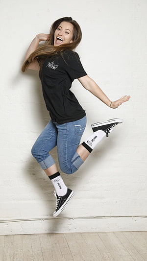 Dani Daniels in Black T-Shirt and Cuffed Denim Jeans Jumping Against White Brick Wall HD phone wallpaper