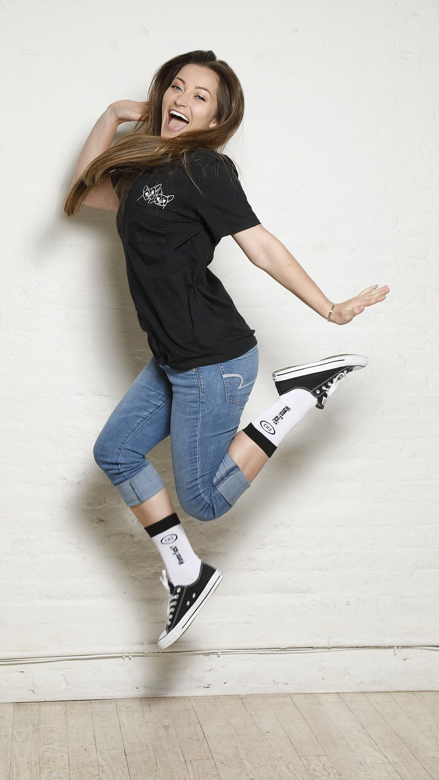 Dani Daniels in Black T-Shirt and Cuffed Denim Jeans Jumping Against White Brick Wall HD phone wallpaper
