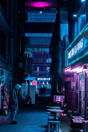 Neon-lit alleyway at night with a hooded figure walking past glowing signs and outdoor seating HD phone wallpaper