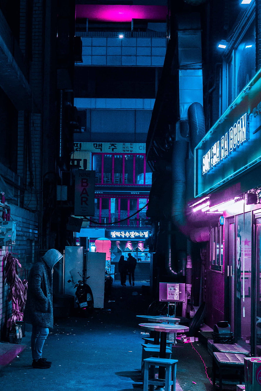 Neon-lit alleyway at night with a hooded figure walking past glowing signs and outdoor seating HD phone wallpaper