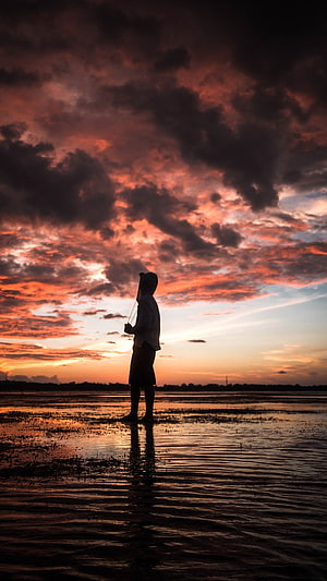 Silhouette of a person standing in shallow water under a dramatic sunset sky with vibrant orange and purple clouds HD phone wallpaper