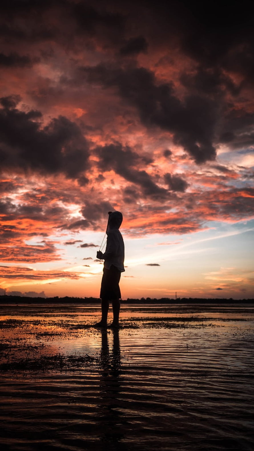 Silhouette of a person standing in shallow water under a dramatic sunset sky with vibrant orange and purple clouds HD phone wallpaper