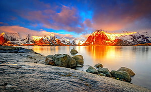 Golden sunset illuminating snow-capped mountains reflected in a calm alpine lake with rocky shoreline HD wallpaper