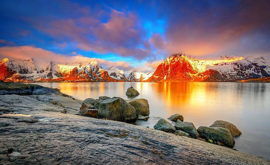 Golden sunset illuminating snow-capped mountains reflected in a calm alpine lake with rocky shoreline HD wallpaper