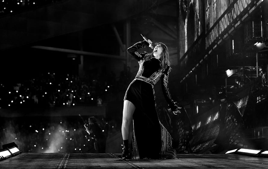 Taylor Swift performing live on stage in a black gown with high slit, singing passionately into a microphone during a concert HD wallpaper