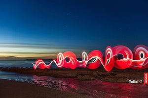 Abstract red light painting swirls over a rocky beach at twilight with a starry night sky HD wallpaper