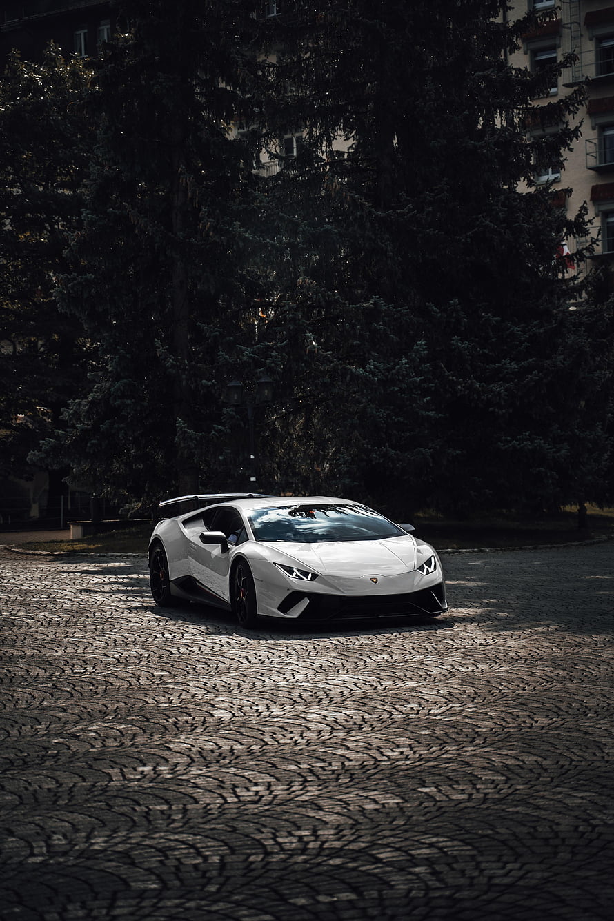 White Lamborghini Aventador SVJ parked on cobblestone street with dark trees and apartment building backdrop HD phone wallpaper
