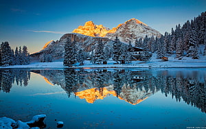 Golden alpenglow on snow-capped mountain peaks reflected in a serene alpine lake surrounded by a winter forest HD wallpaper
