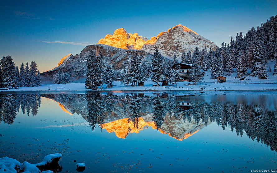 Golden alpenglow on snow-capped mountain peaks reflected in a serene alpine lake surrounded by a winter forest HD wallpaper