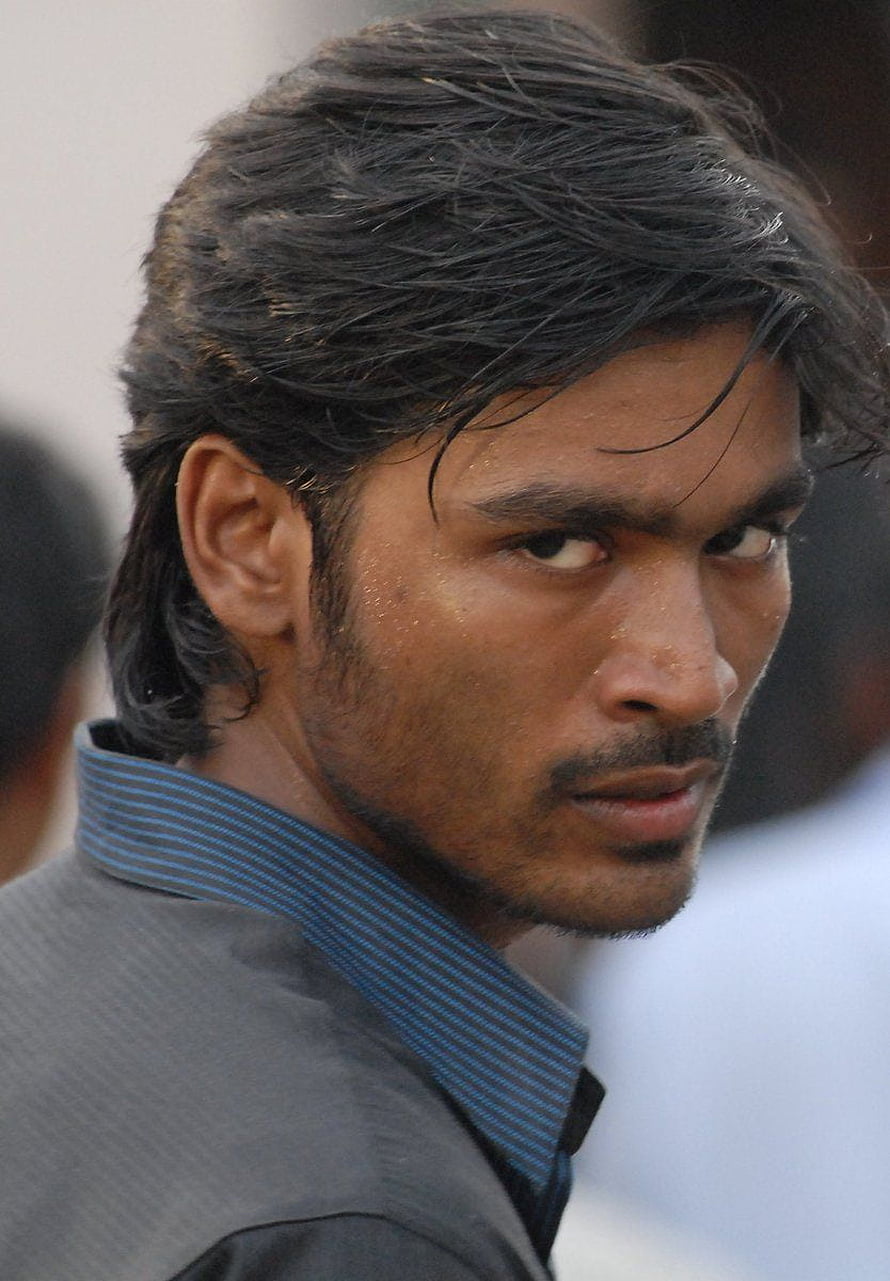 Dhanush in a grey suit with a blue striped shirt collar, looking over his shoulder with a serious expression HD phone wallpaper