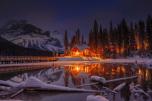 Cozy illuminated cabin by a frozen lake with snowy mountains and pine forest reflection at twilight HD wallpaper