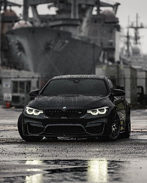 Black BMW M4 Competition parked on wet pavement with naval ship background HD phone wallpaper