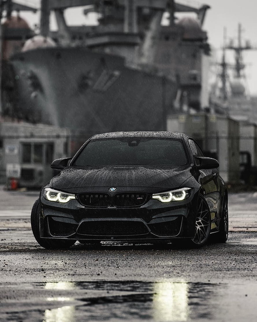 Black BMW M4 Competition parked on wet pavement with naval ship background HD phone wallpaper