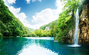 Serene turquoise lake with a cascading waterfall surrounded by lush green forest and blue sky HD wallpaper