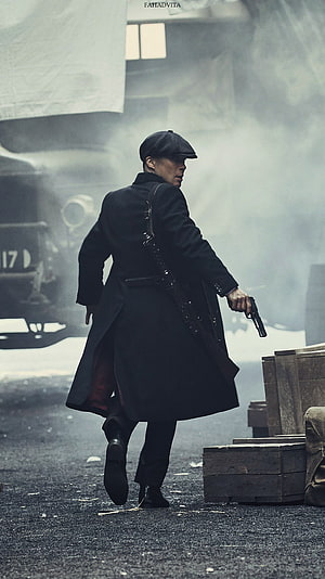 Thomas Shelby from Peaky Blinders in a long black coat and flat cap holding a pistol in a smoky industrial setting HD phone wallpaper