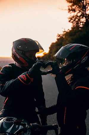 Two motorcyclists in full gear form a heart shape with gloved hands against a golden sunset sky HD phone wallpaper