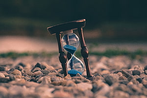 Vintage wooden hourglass with blue sand resting on a bed of smooth river pebbles HD wallpaper