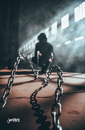Silhouette of a hooded figure crouching behind heavy chains in a dark, smoky industrial corridor HD phone wallpaper